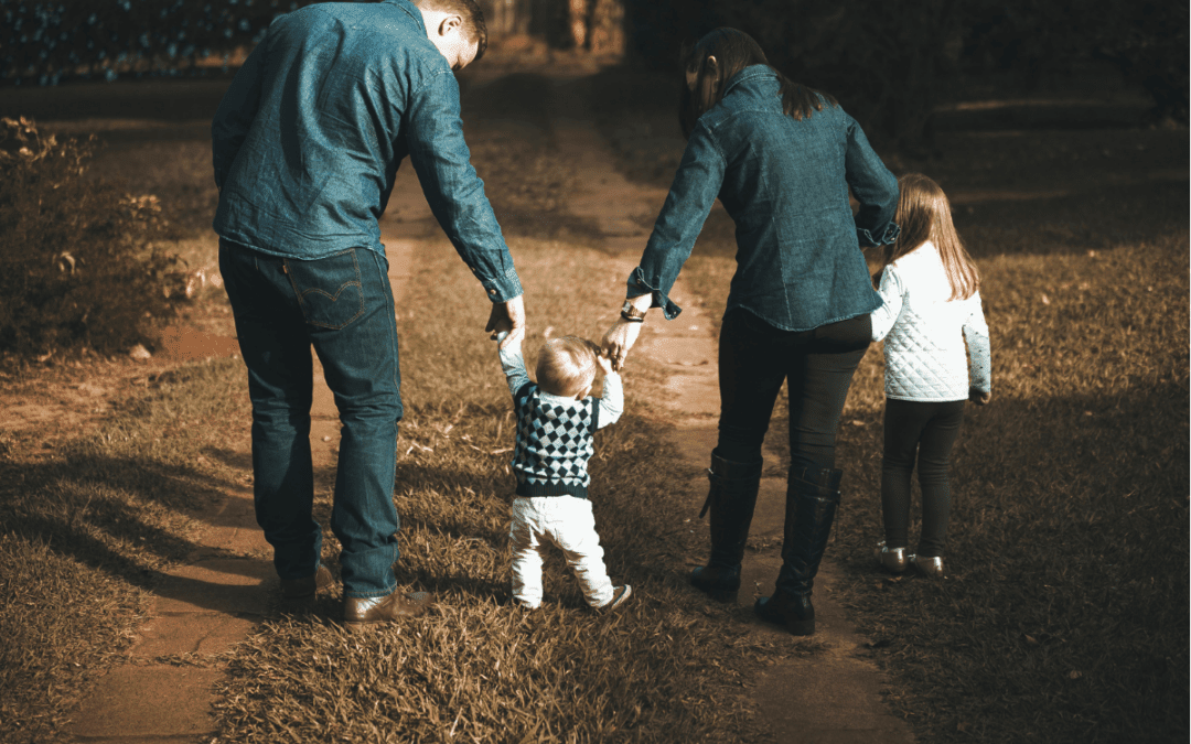A family of four, seen from behind, walks on a sunlit path in a park. Two adults—planning for health insurance over 50—hold a young child’s hands as they help him walk, while an older child walks slightly ahead. Everyone is wearing jeans and jackets.