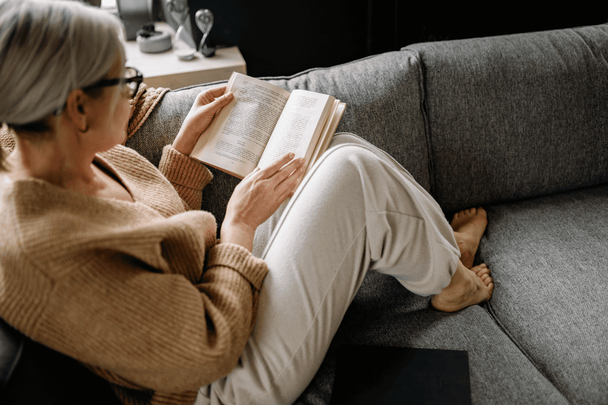 A woman with grey hair and glasses, wearing a beige jumper and light trousers, is relaxing on a grey sofa while reading about health insurance over 50. She sits comfortably with her legs up, focused on the pages. A woman with grey hair and glasses, wearing a beige jumper and light trousers, is relaxing on a grey sofa while reading about health insurance over 50. She sits comfortably with her legs up, focused on the pages of her book which is talking about 5 Essential Questions Over 50s should ask about health insurance