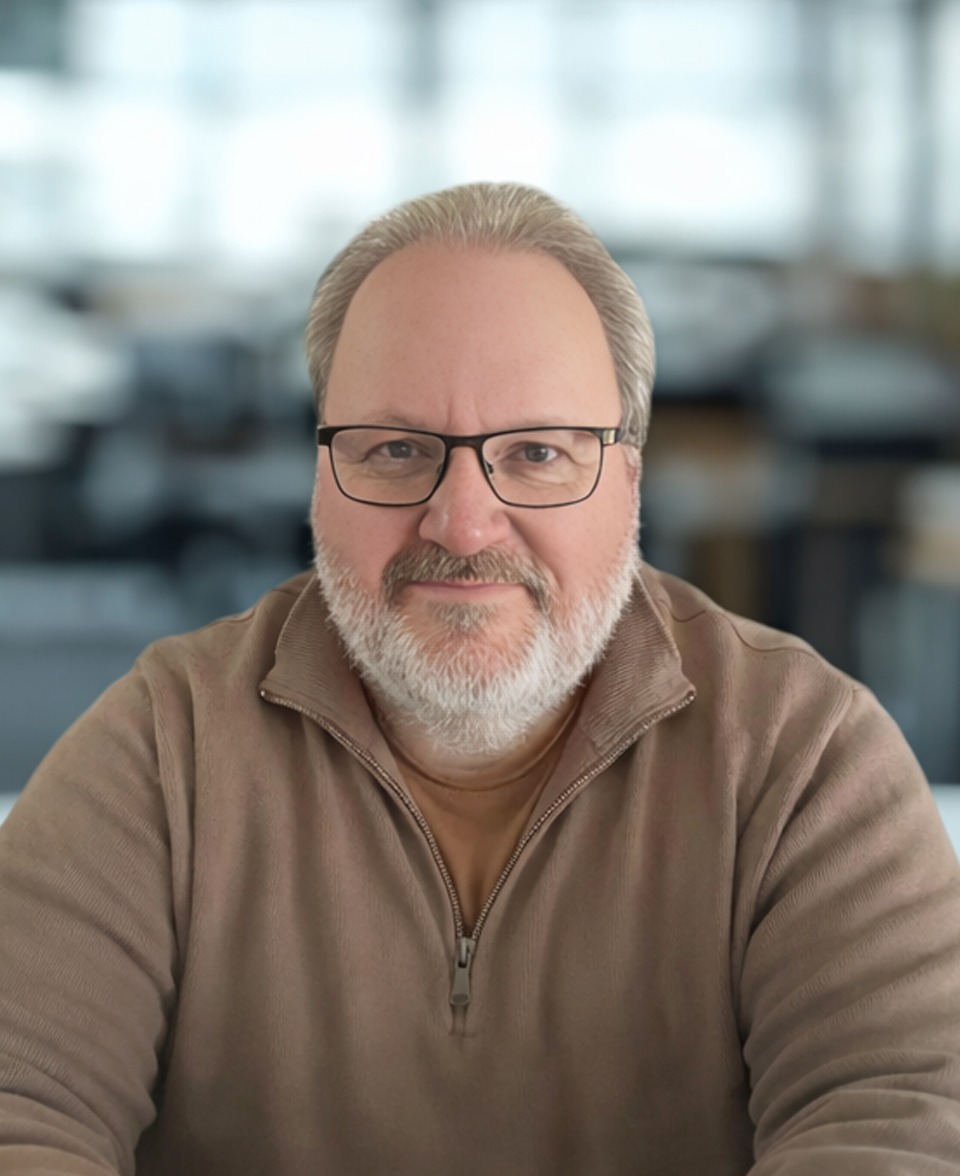 Nigel A middle-aged man with glasses, grey hair, and a beard, wearing a beige zip-up jumper, sits at a table in a bright, blurred office setting.