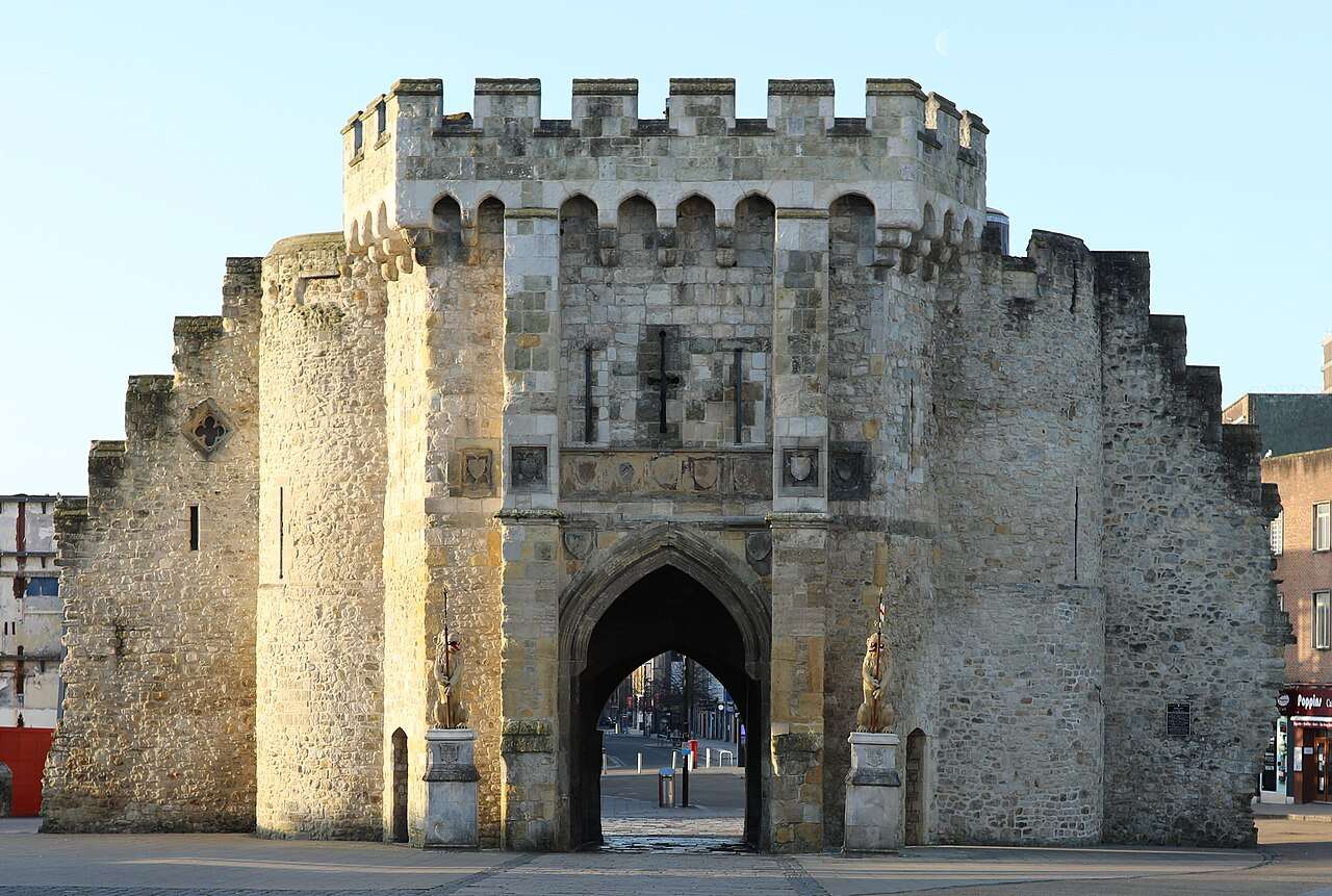 A large stone medieval gatehouse with an arched entrance, battlements on top, and two statues flanking the entryway. The structure stands alone in a paved square with clear sky in the background.