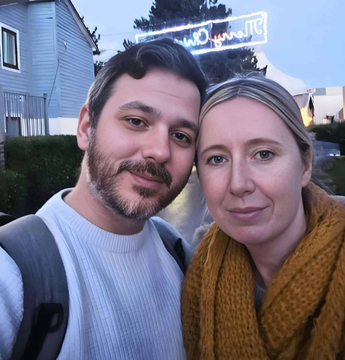 Two people pose closely together outdoors, both looking at the camera. The man has short dark hair and a beard, wearing a white jumper. The woman has blonde hair, tied back, and is wearing a mustard yellow scarf. A neon sign glows in the background.