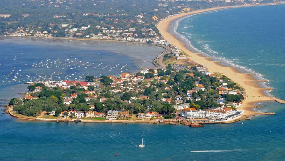 Aerial view of a coastal town with a sandy peninsula, boats in the bay, and buildings surrounded by greenery—bordered by blue ocean water—in picturesque Dorchester, where health insurance options provide peace of mind for residents.