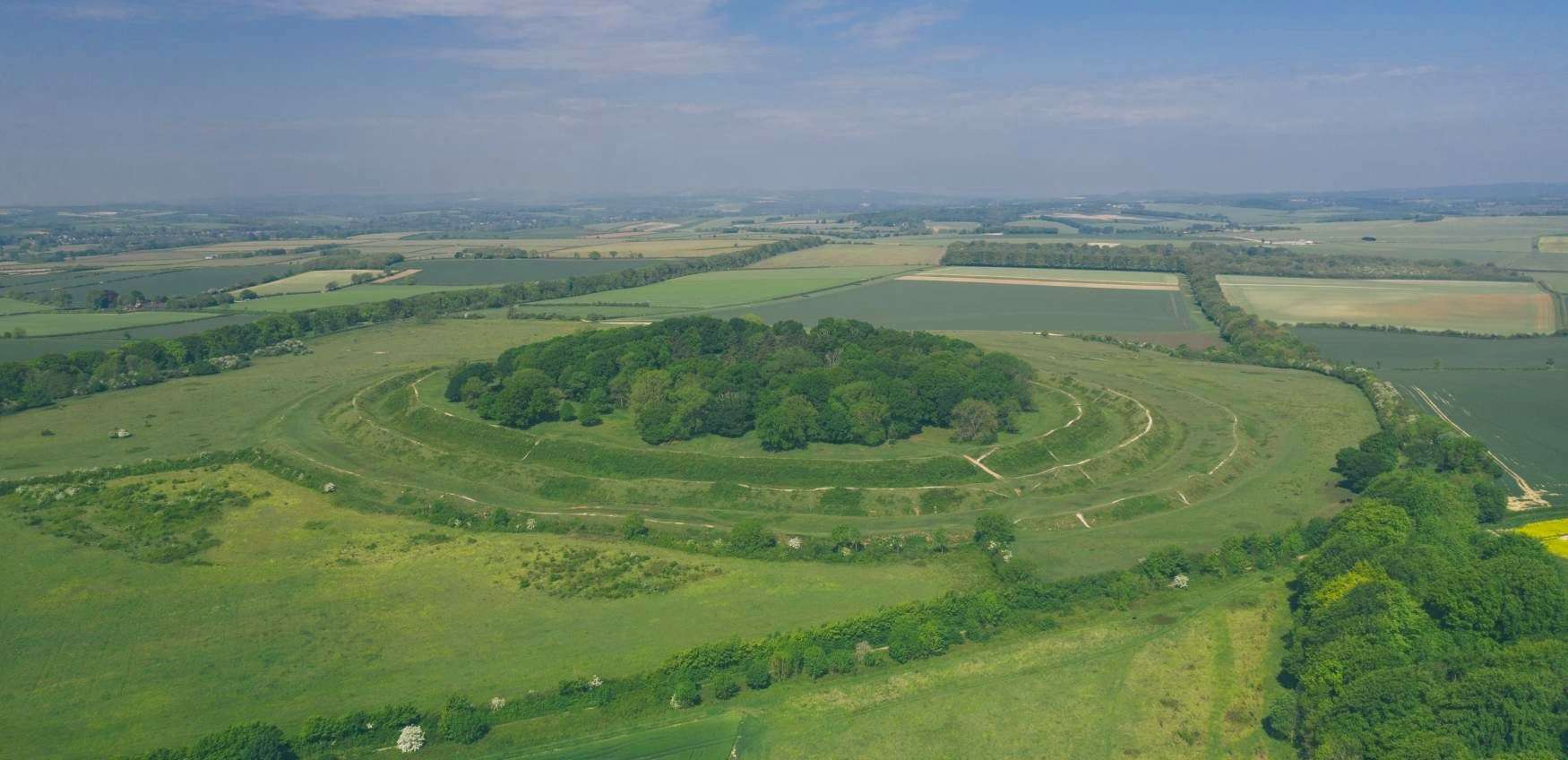 Aerial view of a large ancient circular earthwork surrounded by green fields and trees under a blue sky, with farmland in the distance.