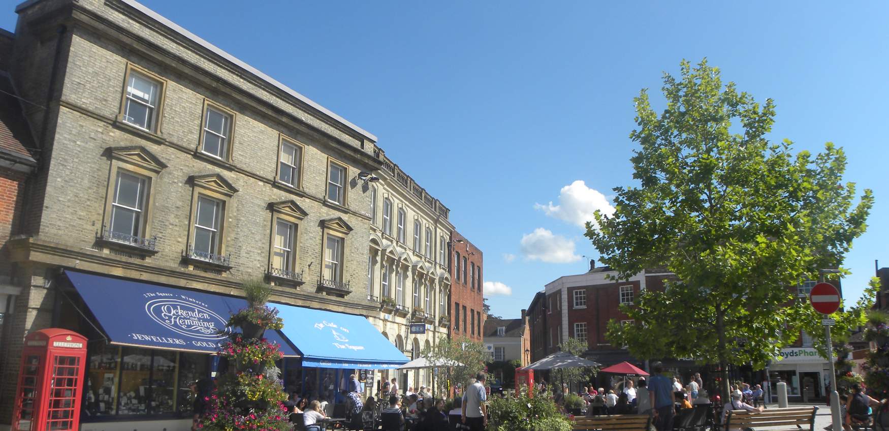 A busy street scene with people sitting outside a café, a large tree, historic buildings, a red telephone box, and a bright blue sky with a few clouds.
