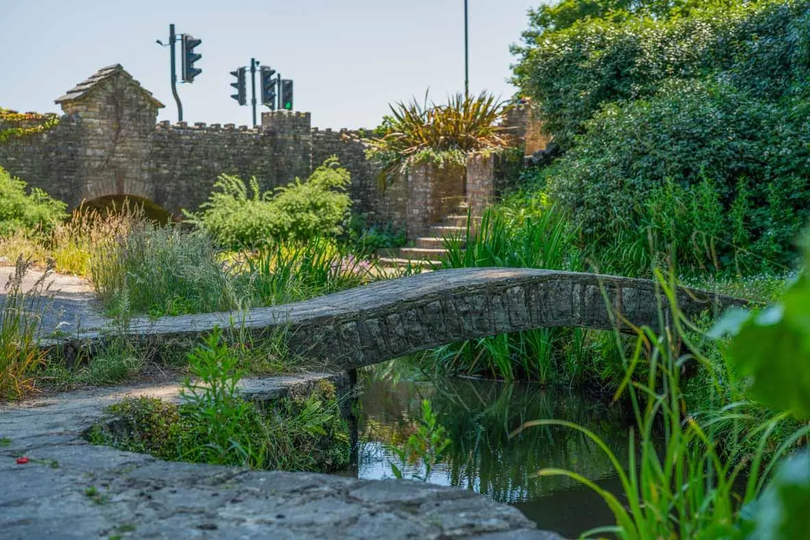 A small stone footbridge crosses a narrow stream in a lush garden, with a stone wall and traffic lights visible in the background.