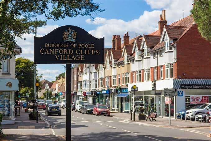 A street view of Canford Cliffs Village in Poole, showing a black village sign, parked cars, shops, and terraced houses under a blue sky with scattered clouds.