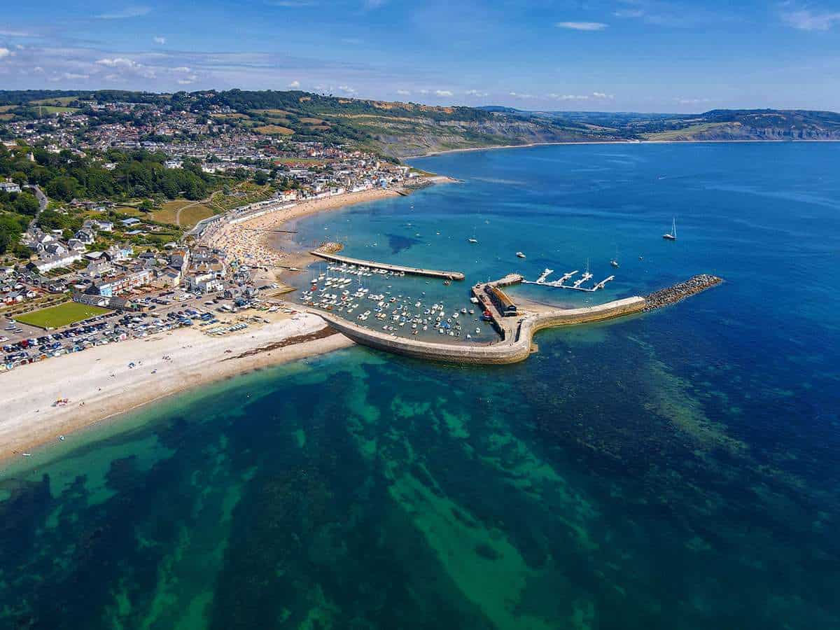 Aerial view of a coastal town with a curved harbour, sandy beaches, clear blue-green water, boats, and distant hills under a blue sky with scattered clouds.
