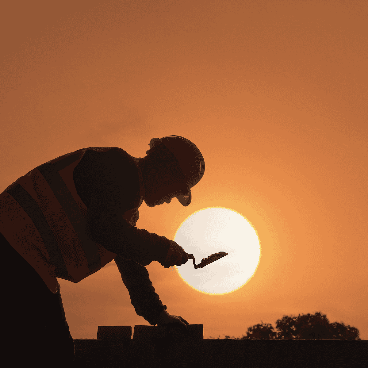 Silhouette of a construction worker in a hard hat and safety vest laying bricks with a trowel against a sunset.